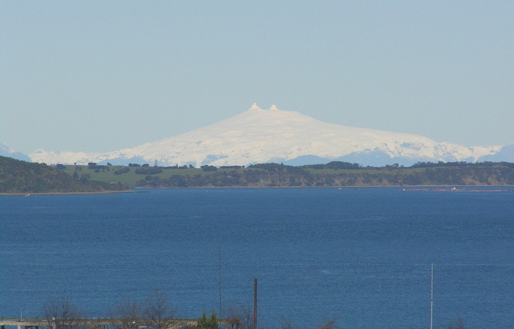 Glacier-clad Melimoyu volcano is seen from the NW from the town of Quellon on the island of Chiloe.  The volcano lies across the Gulf of Corcovado beyond the small island cutting across the image in the foreground.  Two prominent horns at the summit of the volcano rise above the rim of the summit crater.  The large stratovolcano has an 8-km-wide, largely buried ice-filled caldera that is drained by a glacier through a notch in the NE caldera rim.  Two late-Holocene tephra layers have been documented from Melimoyu. Photo by Bryan Freeman, 2005.