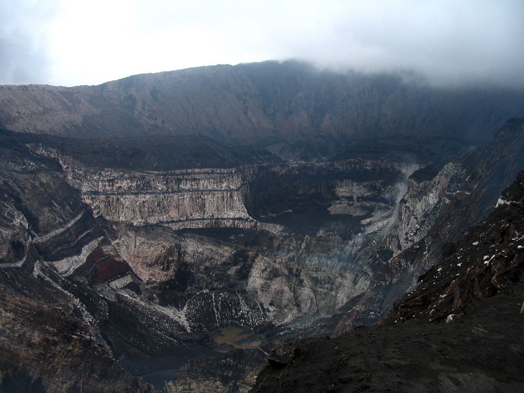 The steep-walled craters of the Marum cone complex at the summit of Ambrym volcano, seen from the east. Ambrym contains a 12-km-wide caldera and is one of the most active volcanoes of the New Hebrides arc. A thick pyroclastic sequence, initially dacitic, then basaltic, overlies lava flows of a pre-caldera shield volcano. Post-caldera eruptions, primarily from Marum and Benbow cones, have partially filled the caldera floor and produced lava flows that ponded on the caldera floor or overflowed through gaps in the caldera rim. Photo by Karoly Nemeth, 2005 (Massey University).