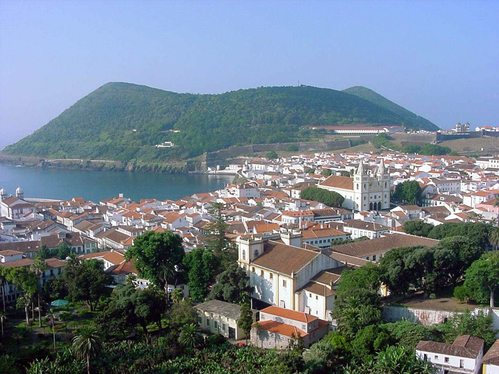 The Monte Brasil pyroclastic cone is seen here beyond the city of Angra do Heroísmo on the south-central coast of Terceira Island. The 1.4 km wide cone is attached to the island by a small peninsula, and is related to the activity of the Guilherme Moniz volcano, which occupies the center of Terceira. Photo by Luís A. da Silveira, 2007 (Wikimedia Commons).