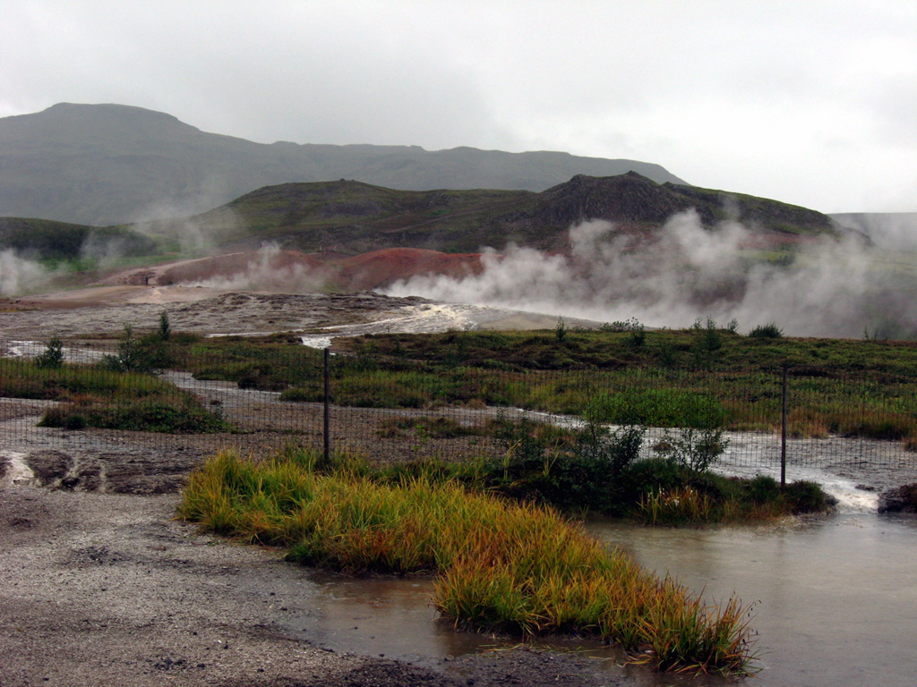 Steam rises from the Geysir geothermal area with the rhyolitic Laugarfjall lava dome in the middle ground behind the mounds of reddish hydrothermally altered clays. The basaltic Bjarnarfjell complex lies on the horizon. The Geysir volcanic system lends its name to geysers around the world and is one of the most frequently visited geological sites in Iceland. No eruptions are known from this volcanic system during the Holocene, although geothermal activity continues and the Strokkur geyser is in almost continuous activity. Photo by Lee Siebert, 2008 (Smithsonian Institution).