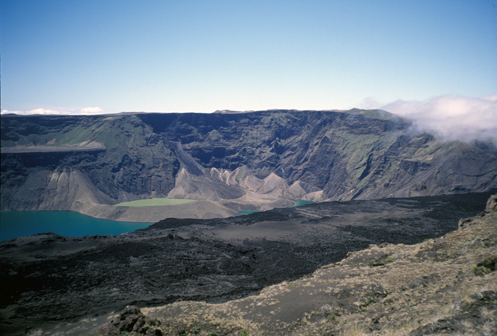 A view from the west caldera rim of Cerro Azul shows a caldera bench covered with young lava flows in the foreground and part of the blue caldera floor lake and a tuff cone filled with greenish lake water.  The photo was taken in July 1998, shortly before an eruption in September 1998 from vents on the south caldera bench and the west caldera floor. Photo by Michael Lang, 1998 (Smithsonian Institution).