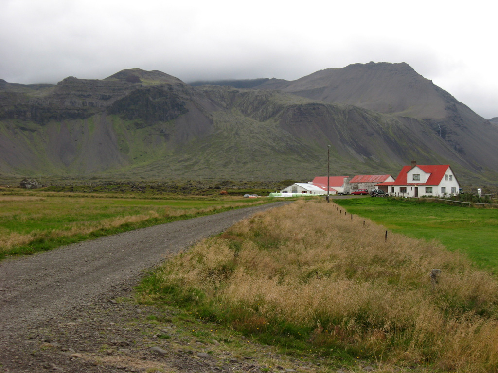 The Bláfeldarhraun lava flow front is seen to the left of the farmhouses. It originated from the cone to the upper left and descended the slope in the center of this view from the south. The flow is one of several Holocene lava flows of the Helgrindur (Lýsuskarð) volcanic system on the Snæfellsnes Peninsula.  Photo by Lee Siebert, 2008 (Smithsonian Institution).