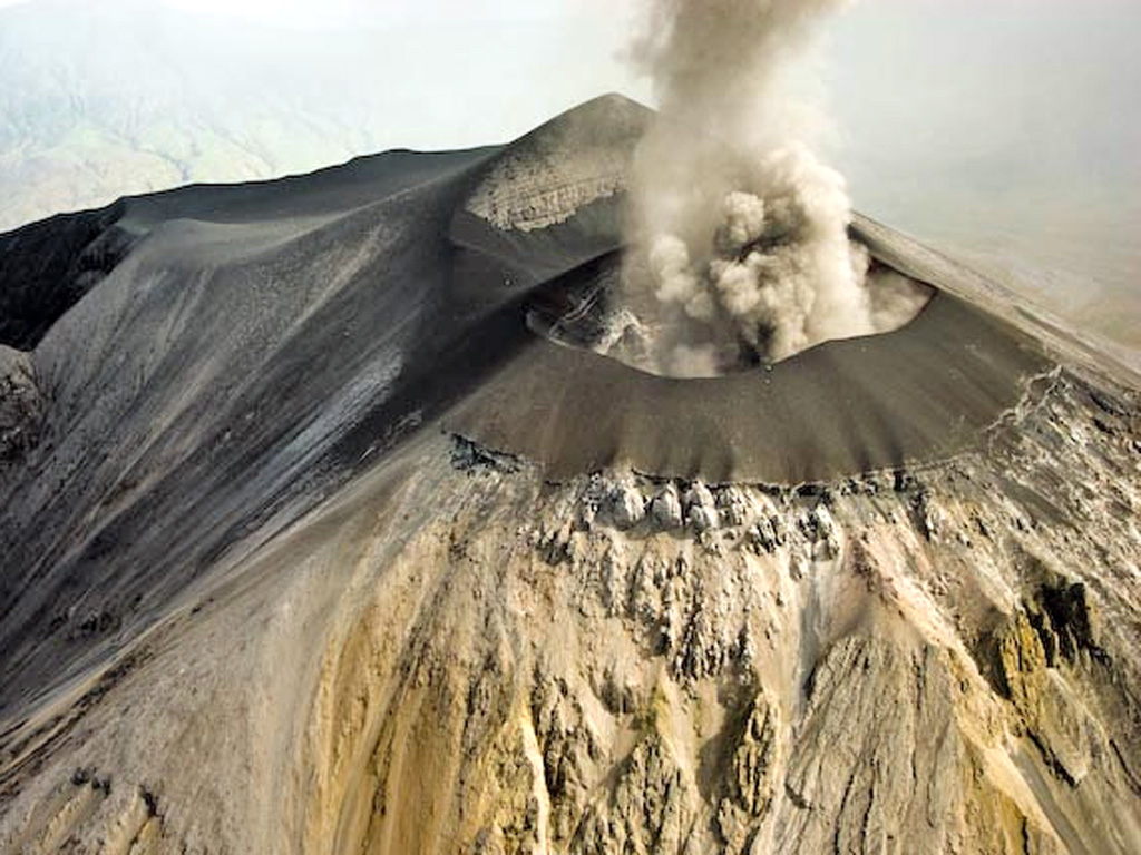 Ash eruption from Ol Doinyo Lengai seen on 12 March 2008 from the NNE. This image shows that the E, N, and W flanks of the cone had buried the original crater rim. Oversteepening of the cone flank in places resulted in small landslides which can be seen just below the cone as dark material covering the lighter areas of older weathered carbonatite. The peak beyond the ash plume is the summit. Photo by Benoît Wilhelmi, 2008.