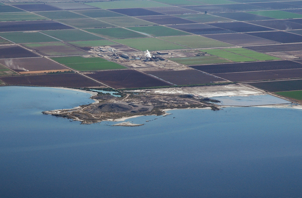 Obsidian Butte on the SE shore of the Salton Sea is seen in an aerial view from the NW.  Obsidian Butte is one of five small rhyolitic lava domes extruded onto Quaternary sediments of the Colorado River delta; the summit of the dome lies 40 m below sea level.  Two domes, Mullet Island and Red Island (not visible in this view), form small islands just offshore to the NE of Obsidian Butte.  A steam plume rises from the Salton Sea geothermal field behind the dome.   Photo by Bruce Perry, 2005 (California State University Long Beach).