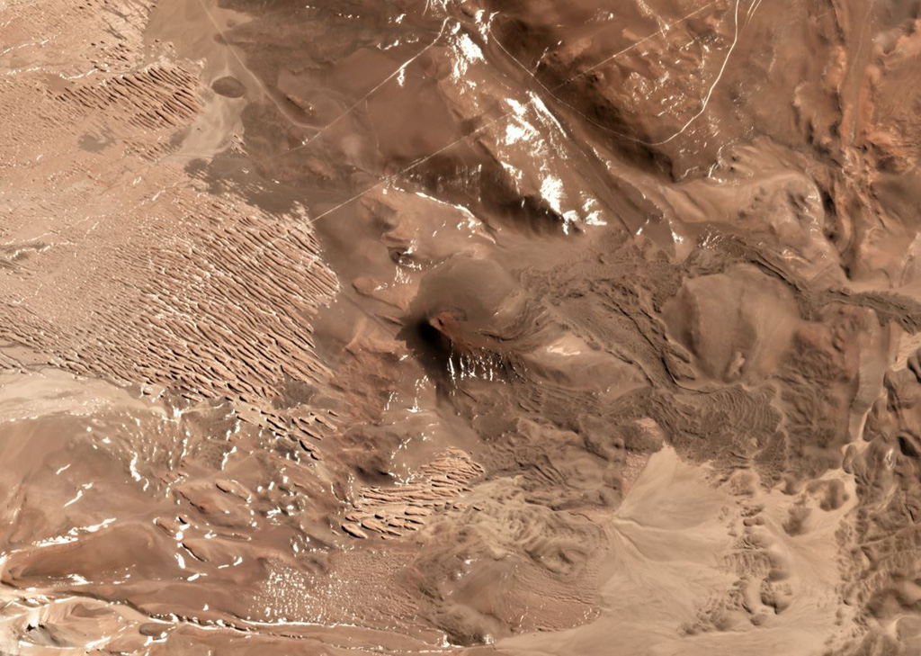 This unnamed scoria cone near the Chile/Argentina border has produced lava flows to the east, visible in this July 2019 Planet Labs satellite image Monthly Mosaic (N is at the top; this image is approximately 8.5 km across). The visible flows have clear lobate or broader flow boundaries, levees, and pressure ridges, while other lavas have been buried by sand deposits. Satellite image courtesy of Planet Labs Inc., 2019 (https://www.planet.com/).
