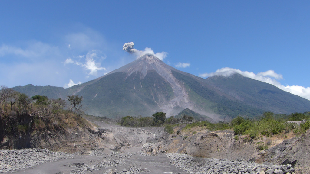 An ash plume from a small explosion at Fuego is being dispersed towards the E by the wind in this 8 March 2020 photo taken from the new Las Lajas bridge. A new bridge was constructed after the previous one was destroyed by the 3 June 2018 eruption that devastated nearby areas. The ravine in the foreground is Barranca Las Lajas, which channeled the majority of pyroclastic flow material produced in the 3 June eruption. Deposits from these pyroclastic flows are visible in the foreground, with dead trees and scorched vegetation on the left margin of the ravine showing some of the impacts of the hot flow. Photo by Ailsa Naismith, 2020.