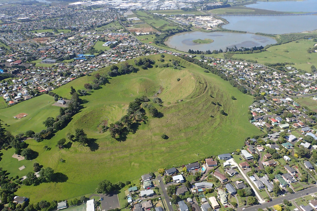 Mangere Mountain is a monogenetic cone within the Auckland Volcanic Field with spatter and scoria components, and several craters with extensive lava flows. The Mangere Lagoon tuff ring is to the upper right in this 2018 view from the NE. Photo by Bruce Hayward, 2018.