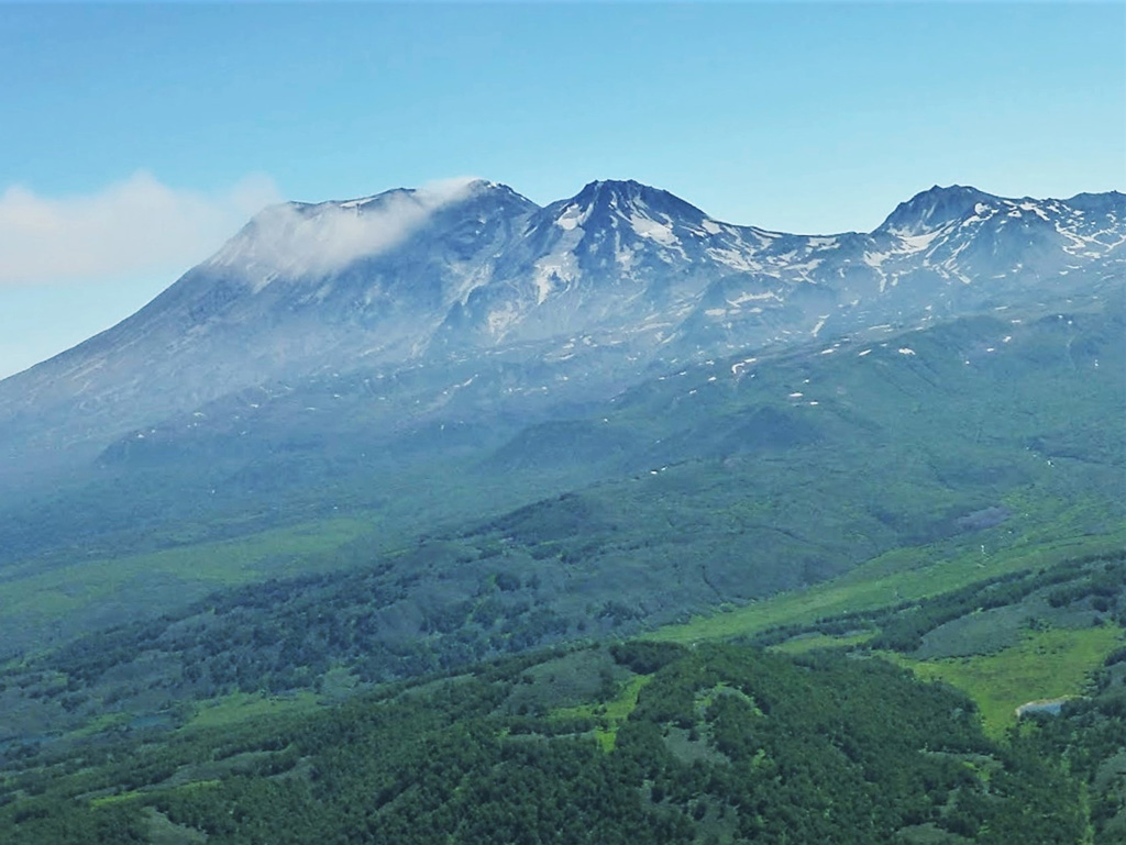 Zhupanovsky has four large edifices that form a WNW-trending ridge, with the active Priemysh cone emitting a gas plume towards the N, which was then redispersed to the W across this image. The older Zhupanovsky peak is to the left and the Bastion edifice is to the right. Photo by Janine Krippner, 2014.