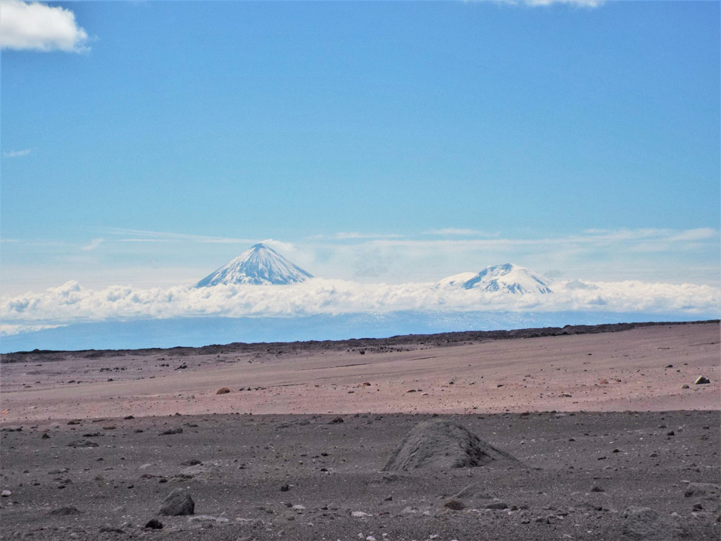 This July 2015 photo looks across the 2010 Sheveluch block-and-ash flow towards the 1964 debris avalanche deposit. Klyuchevskoy (left) and Ushkovsky (right) are in the background, with a gas plume emitting from the former. The section of deposit in the foreground is darker because of cloud cover. Photo by Janine Krippner, 2015.