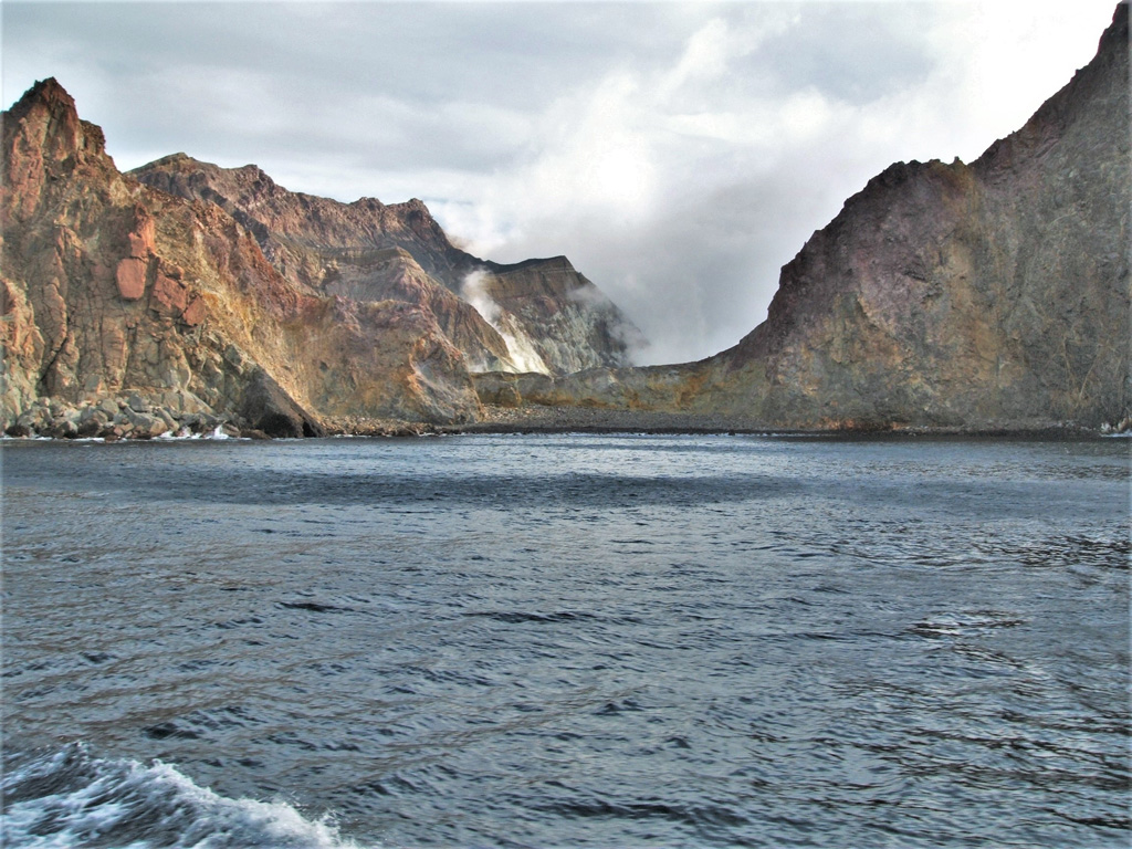 This August 2005 view is looking into the Whakaari/White Island crater through Wilson Bay at the SE of the island. Steam and gases are rising from the crater lake at the far end of the crater, as well as from fumaroles along the crater rim. The units exposed in the inner and outer walls are variably weathered and oxidized lavas, tuffs, and breccias. Photo by Janine Krippner, 2005.