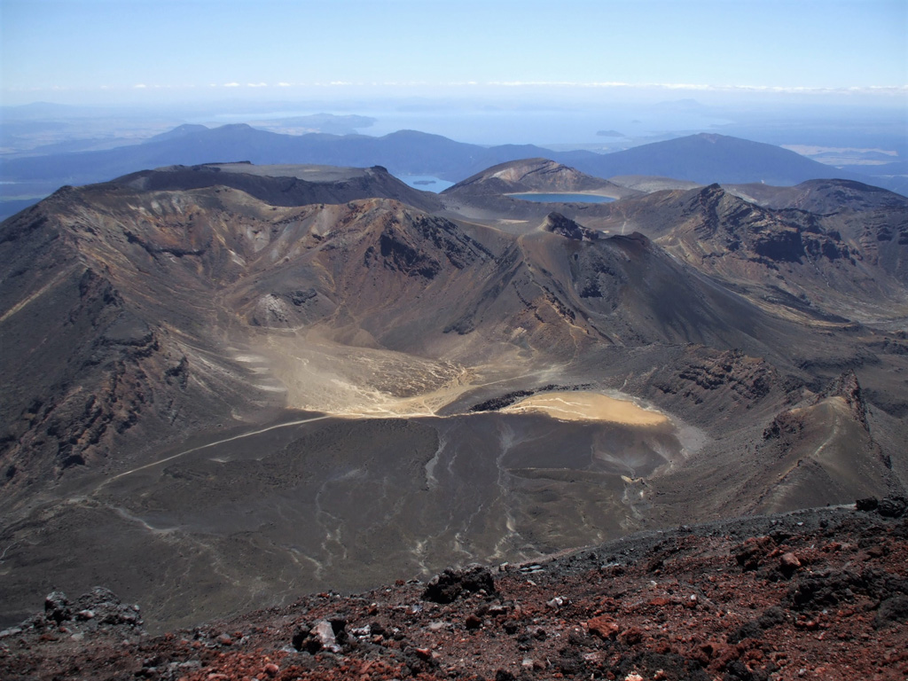 This 2008 view from the Ngauruhoe summit (red scoria at the bottom) looks across Tongariro and to Lake Taupō in the distance. Within Tongariro going in a clockwise direction, South Crater is in the foreground, North Crater is to the upper left, Blue Lake is to its right, and Red Crater is below it. Photo by Janine Krippner, 2008.